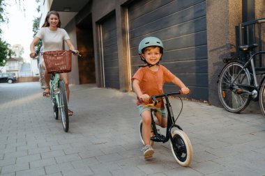 Kask takan oyuncu bir çocuk ve annesi güneşli bir sokakta bisiklet sürerken mutlu bir şekilde eğleniyor..