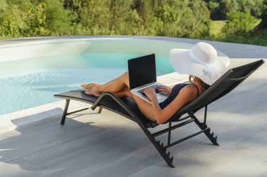 Beyaz şapkalı bir kadın havuz kenarındaki şezlongda dizüstü bilgisayarıyla çalışıyor. Uzaktan kumandalı çalışma, boş zaman ve açık havada rahatlama dengesi. Metin için boşluk