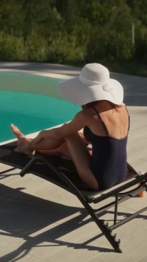 Mayo giymiş ve geniş kenarlı beyaz şapkalı bir kadın güneş kremi sürüyor. Açık havuz kenarında bir şezlongda otururken dinleniyor. Güneş koruması