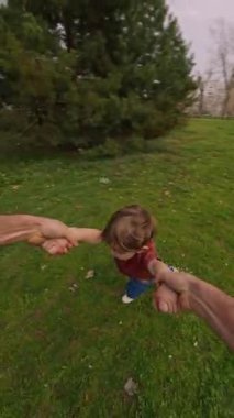 Anne ve baba, mutlu gülen çocuklarının ellerini tutup onları parkta bir çember etrafında döndürüyor. POV görüntüsü, babası neşeli oğluyla dışarıda oynuyor. Mutlu çocukluk kavramı. Dikey video