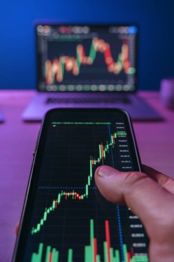 POV görünümü, akıllı telefon tutan tüccar, kripto ve borsa eğilimlerini analiz ederek ticaret uygulaması kullanarak neon ışıklı ofisinde arka planda birden fazla monitör ile birlikte. 