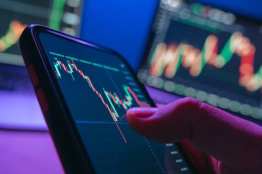 Şifreleme ve borsa eğilimlerini analiz ederek arka planda birden fazla monitörle neon ışıklı ofisinde ticaret uygulaması kullanarak akıllı telefonu tutan tüccarın elini kapat.