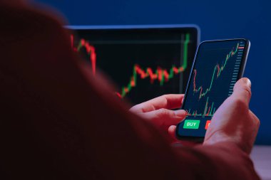 Akıllı telefonu tutan tüccar elleri, finansal, borsa ve kripto para birimi verilerini analiz etmek için ticaret uygulaması kullanıyor, ekranda kırmızı ve yeşil şamdan çizelgeleri bulunuyor..