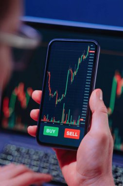 Arkaplanda dizüstü bilgisayarda sergilenen borsa verileriyle akıllı bir telefon üzerinden kripto para birimi grafiklerini yakından inceleyen bir tüccarın omuz hizasında..