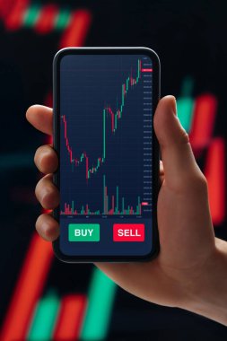 Kapat, borsa uygulamalı akıllı telefonu elinde tutan tüccar arka planda şamdan diyagramına karşı. Çevrimiçi ticaret ve yatırım kavramı