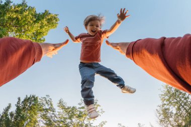 Güneşli bir günde gökyüzü ve ağaçlarla kaplı bir ebeveyn tarafından terk edilirken neşeli bir şekilde gülümseyen heyecanlı genç çocuk. POV bakış açısı, bir babanın yüksekten atması ve gülen oğlunu parkta yakalaması..