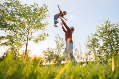 Neşeli bir baba, günbatımında ağaçlar ve taze yeşil çimlerle çevrili güneşli bir parkta oynayarak küçük oğlunu havaya fırlatıyor. Babalar Günü ve aile bağı kavramı. Boşluğu kopyala