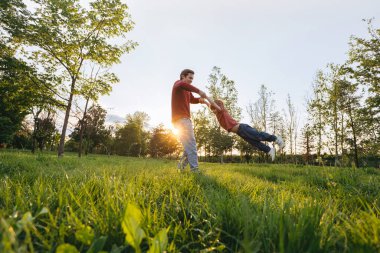 Neşeli genç baba gün batımında mutlu oğlunu çimenli bir parkta döndürüyor. Gülümseyen baba dışarıda neşeli çocuğuyla oynuyor. Aile bağları ve bağları. Metin için boşluk