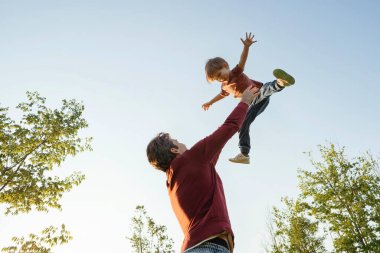 Baba ve oğul parkta birlikte oynuyorlar, baba gülücük saçan oğlunu havaya fırlatıyor, eğleniyor ve ailece vakit geçiriyorlar. Mutlu çocukluklar ya da Babalar Günü konsepti. Metin için boşluk