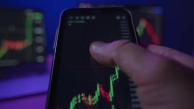 Birinci şahıs görüşü, borsa ve kripto para birimini analiz etmek ve satın almak için telefonunu kaldıran tüccar masasının önünde gece kripto çizelgeleri sergilerken bir ticaret uygulaması kullanıyor.