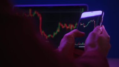 Akıllı telefonu tutan tüccar elleri, finansal, borsa ve kripto para birimi verilerini analiz etmek için ticaret uygulaması kullanıyor, ekranda kırmızı ve yeşil şamdan çizelgeleri var, koyu neon ışıklı ofis ofisinde..