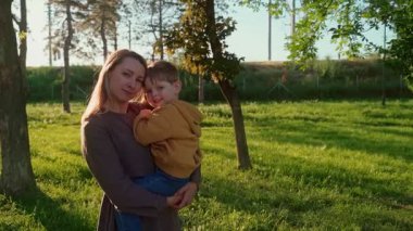 Ağaçlar ve doğa ile çevrili güneşli, yeşil bir parkta neşeli oğlunu kucaklayan bir annenin şefkatli anı. Anne sevimli oğlunu parkta tutuyor ve gülümsüyor. Mutlu anneler günü konsepti
