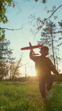 Gülümseyen mutlu genç çocuk gün batımında uzun ağaçlarla çimenli bir tarlada koşuyor, oyuncak bir uçak tutuyor ve dışarıda yaratıcı oyun saatlerinin tadını çıkarıyor, uçma hayalleri kuruyor. Ağır çekim. Dikey video