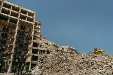 Yıkım bölgesinde bir kazıcıyla yıkılan bir binanın yıkıntıları, yıkıntı ve yıkık betonla çevrili inşaat atıklarını taşıyor. Boşluğu kopyala