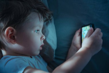 Küçük bir çocuğun gece yatağında yatarken, akıllı telefondan çizgi film izlerken ya da mobil video oyunları oynarken geniş açıyla çekilmiş görüntüleri. Ekran bağımlılığı ve aygıt kullanımı. 