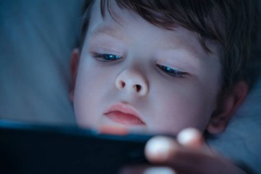 Genç bir çocuğun gece yatağında yatarken, gözleri akıllı telefon ekranına kilitlenirken, mobil oyunlar oynarken ya da çizgi film izlerken. Gadget bağımlılığı ve aşırı kullanımı. 
