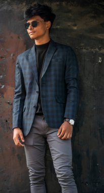 Siyah gri takım elbiseli, güneş gözlüklü, ayakta durup kameraya bakan genç bir çocuk..