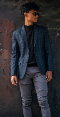 Siyah gri takım elbiseli, güneş gözlüklü, ayakta durup kameraya bakan genç bir çocuk..