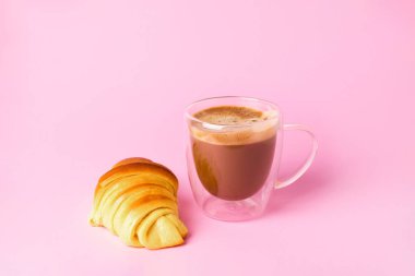 Açık pembe arka planda köpüklü ve lezzetli bir kruvasan ile bir fincan kahve.