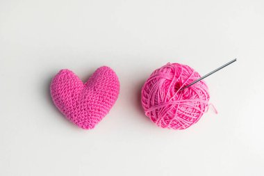 Tığ işi pembe kalp, iplik yumağı ve arka planda örgü kancası, üst manzara.