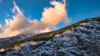 Pine forest on the tope of mountain range and clouds touching mountain peaks, illuminated in sunset light. Ski winter holidays in Andorra, Pyrenees