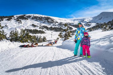 Mother and daughter, skiers, standing on snowy road, Huskey dog sledding, mountains and forest in background. Winter ski holidays, Andorra, Pyrenees