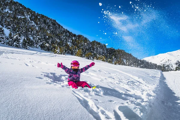 Smiling girl sitting on snow and throwing snow powder up to the air, mountains and forest in a background. Winter ski holidays, Andorra, Pyrenees