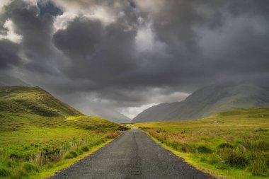 Road leading trough Doolough Valley, between Glenummera and Glencullin mountain ranges illuminated by sunlight with dark dramatic sky, Mayo, Ireland