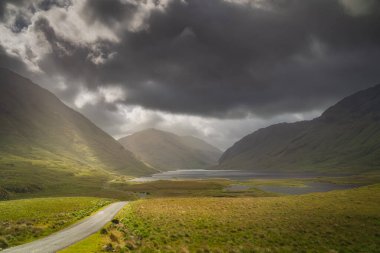 Road leading trough Doolough Valley with lakes, Glenummera and Glencullin mountain ranges illuminated by sunlight, dark dramatic sky, Mayo, Ireland