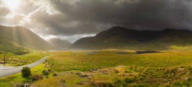Panorama of Doolough Valley with lakes, Glenummera and Glencullin mountain ranges illuminated by sunlight, dark dramatic sky, County Mayo, Ireland