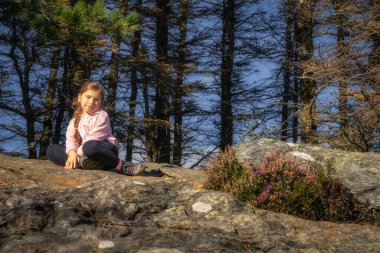 Ormandaki bir kayanın üzerinde oturan, gün ışığıyla aydınlanan genç, gülümseyen kız. Aktif çocuk, Glendalough 'da yürüyüş, Wicklow Dağları, İrlanda