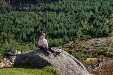 Genç, gülümseyen bir kız, bir kayanın üzerinde oturuyor, arka planda orman ve vadi ile güneş ışığıyla aydınlatılıyor. Wicklow Dağları 'nda yürüyüş, İrlanda