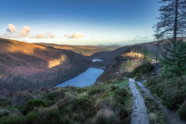 Dağın tepesindeki tahta kaldırımda Glendalough gölleri ve ormanı manzaralı. Wicklow Dağları, İrlanda 'da gün batımında yürüyüş