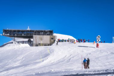 Canlı bir kayak merkezi sahnesinde, güzel tasarlanmış modern bir kulübe var. Etrafı coşkulu kış sporu severlerle çevrili. Temiz mavi gökyüzünün altında, el değmemiş karda eğleniyorlar.