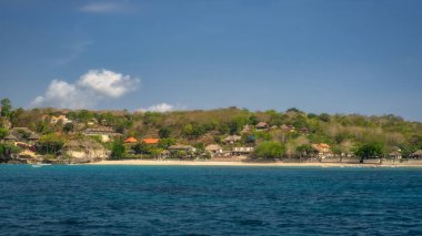 Büyüleyici ve güzel bir plaj sahnesi sergilenmekte, bereketli bir kıyı manzarasının yanında inanılmaz açık mavi suları sergiliyor. Parlak, güneşli bir gökyüzü altında, Nusa Lembongan, Bali, Endonezya