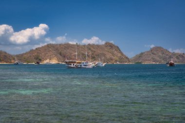 Berrak mavi sularda demirlemiş teknelerin bulunduğu sakin bir sahil manzarası hayal edin, yeşil tepeler ve canlı bir gökyüzü tarafından çerçevelenmiş, doğaya mükemmel bir kaçış, Labuan Bajo, Komodo, Endonezya
