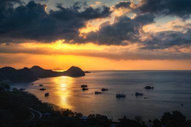 Ufukta büyüleyici bir günbatımı ışıldıyor, sakin denize altın yansımalar yapıyor, kıyıda sallanan kayıklar, sakin bir atmosfer yaratıyor, Labuan Bajo, Komodo, Endonezya