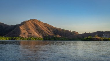 A breathtaking serene view of gentle hills beautifully reflected in the calm waters beneath a clear blue sky, showcasing natures peaceful and idyllic beauty perfectly, Komodo archipelago, Indonesia clipart