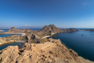 Engebeli dağların engebeli okyanuslarla zarif bir şekilde buluşması, dingin mavi sular, Padar Adası, Pembe Plaj, Komodo, Endonezya ile birlikte nefes kesen kıyı şeritlerinin çarpıcı panoramik manzarası.