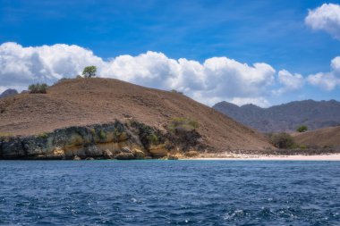 Huzurlu bir kıyı manzarası, dalgalı tepeler, canlı mavi sular ve tüylü bulutlarla kaplı berrak bir gökyüzü, Komodo, Endonezya