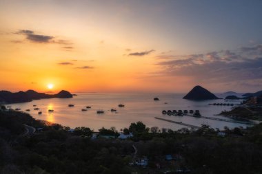 Siluetli adalar ve teknelerin bir araya gelip inanılmaz güzel bir sahil sahnesi oluşturduğu sakin bir deniz boyunca uzanan büyüleyici bir günbatımı manzarasının tadını çıkarın, Labuan Bajo, Komodo, Endonezya