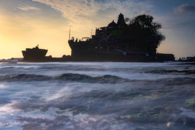 Göz kamaştırıcı bir sahil sahnesinde canlı bir günbatımına karşı siluetli bir Tanah Lot Tapınağı sergileniyor, sakin sular ve dokunulmamış doğa ile çevrili, doğa severler için ideal, Bali, Endonezya