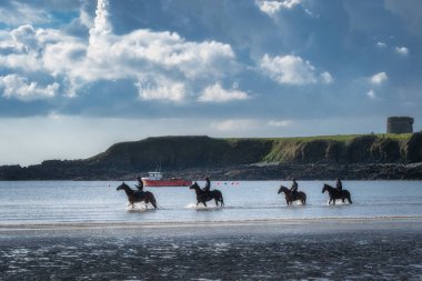 Loughshinny, İrlanda, 1 Mayıs 2021 'de yetenekli biniciler, gökyüzündeki canlı bulutlar tarafından güzelce çerçevelenmiş, ışıldayan sularda zarif bir şekilde at sürerek, herkes için gerçek bir cennet manzarası yarattılar.