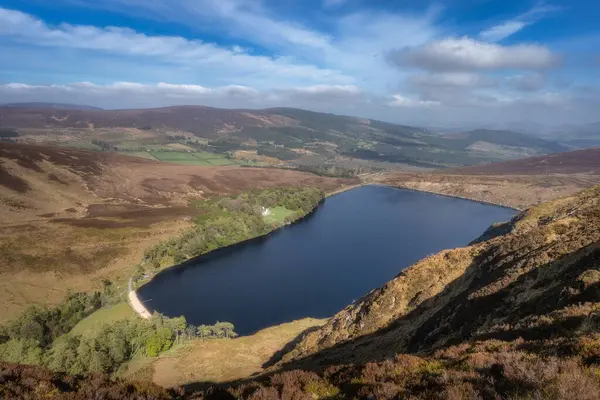 Yuvarlanan tepelerin arasına yerleşmiş sakin Bray Gölü 'nün nefes kesici hava manzarasını deneyimleyin, büyüleyici yemyeşil bir manzara ve yukarıda yüzen dramatik bulutlar, Wicklow, İrlanda