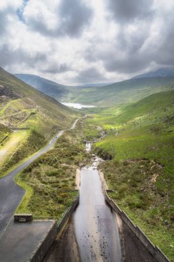 Görkemli yas dağları ve dramatik bulutlu gökyüzü ile çevrili kıvrımlı bir nehir içeren yemyeşil, yemyeşil bir vadi deposunun çarpıcı manzarası, Kuzey İrlanda