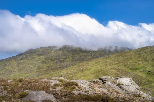 Huzurlu ve sakin Slieve Foye dağ sahnesi sergileniyor. Sergilenen bulutlar, yemyeşil tepeler ve engebeli kayalık arazi üzerinde sürükleniyor. Hepsi geniş, parlak, mavi bir gökyüzü altında, Carlingford, İrlanda.