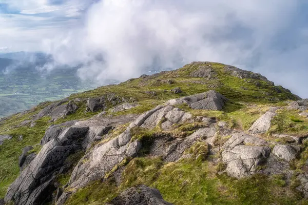 Kayalık Slieve Foye dağının bulutların arasından zarif bir şekilde yükselen büyüleyici ve nefes kesici manzarasını deneyimleyin, canlı yemyeşil ve engebeli doğal güzelliği sergiliyor, Carlingford, İrlanda