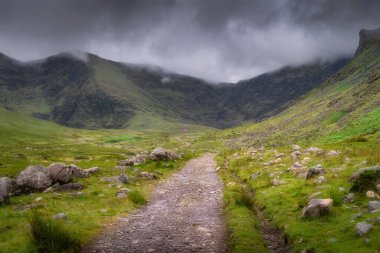 Huzurlu ve huzurlu bir patika, gür, canlı yeşil bir vadi boyunca uzanır, yüksek, görkemli Carrauntoohil dağıyla çevrili dramatik bulutlu gökyüzü altında, Kerry, İrlanda