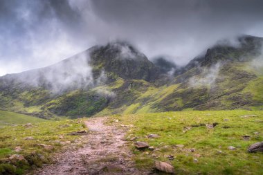 Puslu Carrauntoohil dağının ve kıvrımlı bir yolun olduğu büyüleyici manzaranın tadını çıkarın. Çevresi canlı yeşil çimenler ve yukarıda tüylü bulutlarla çevrili. Kerry, İrlanda.