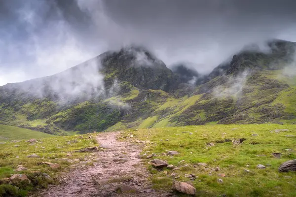 Puslu Carrauntoohil dağının ve kıvrımlı bir yolun olduğu büyüleyici manzaranın tadını çıkarın. Çevresi canlı yeşil çimenler ve yukarıda tüylü bulutlarla çevrili. Kerry, İrlanda.